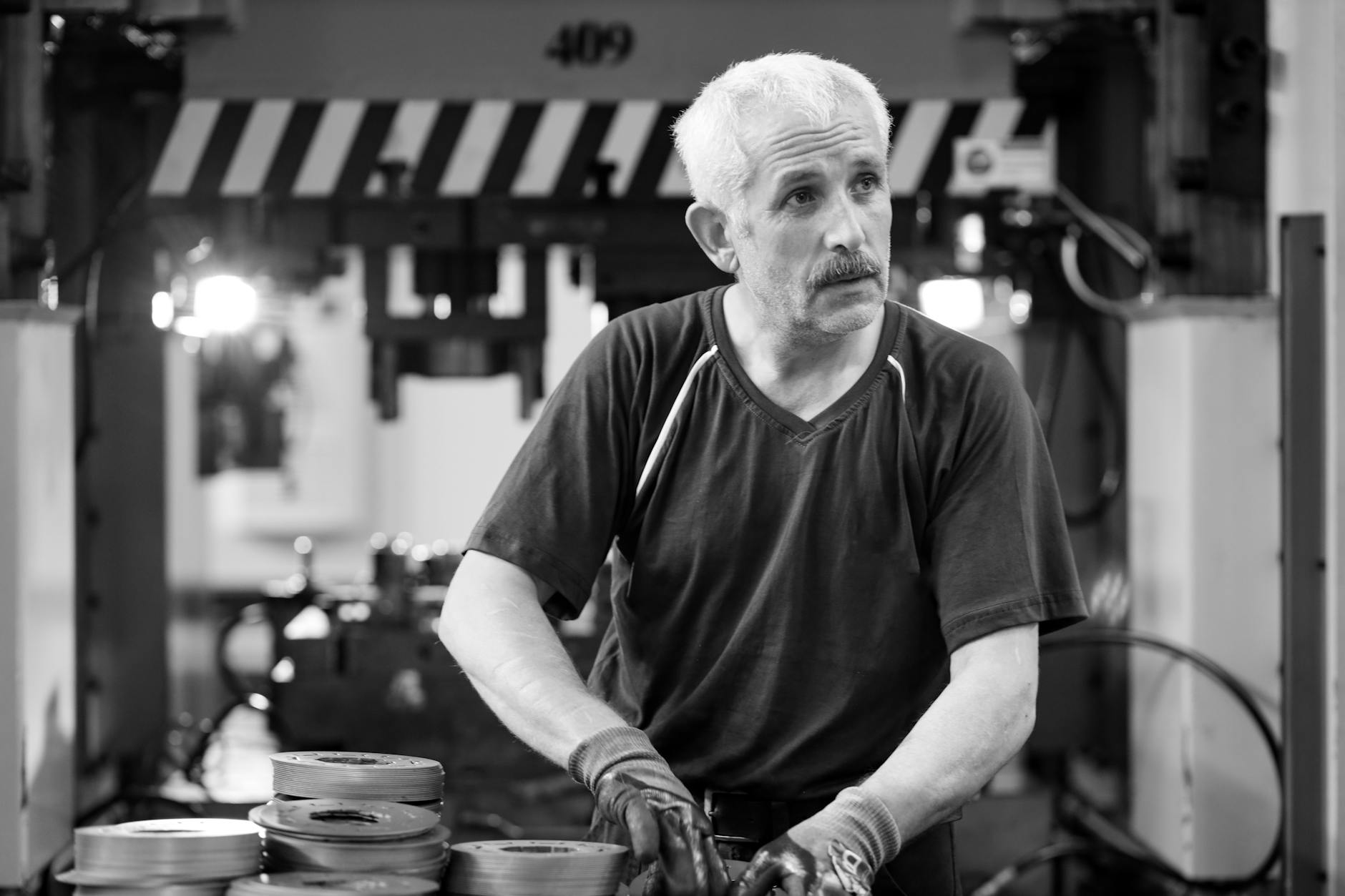 Black and white photo of an elderly worker with gloves in a workshop setting.