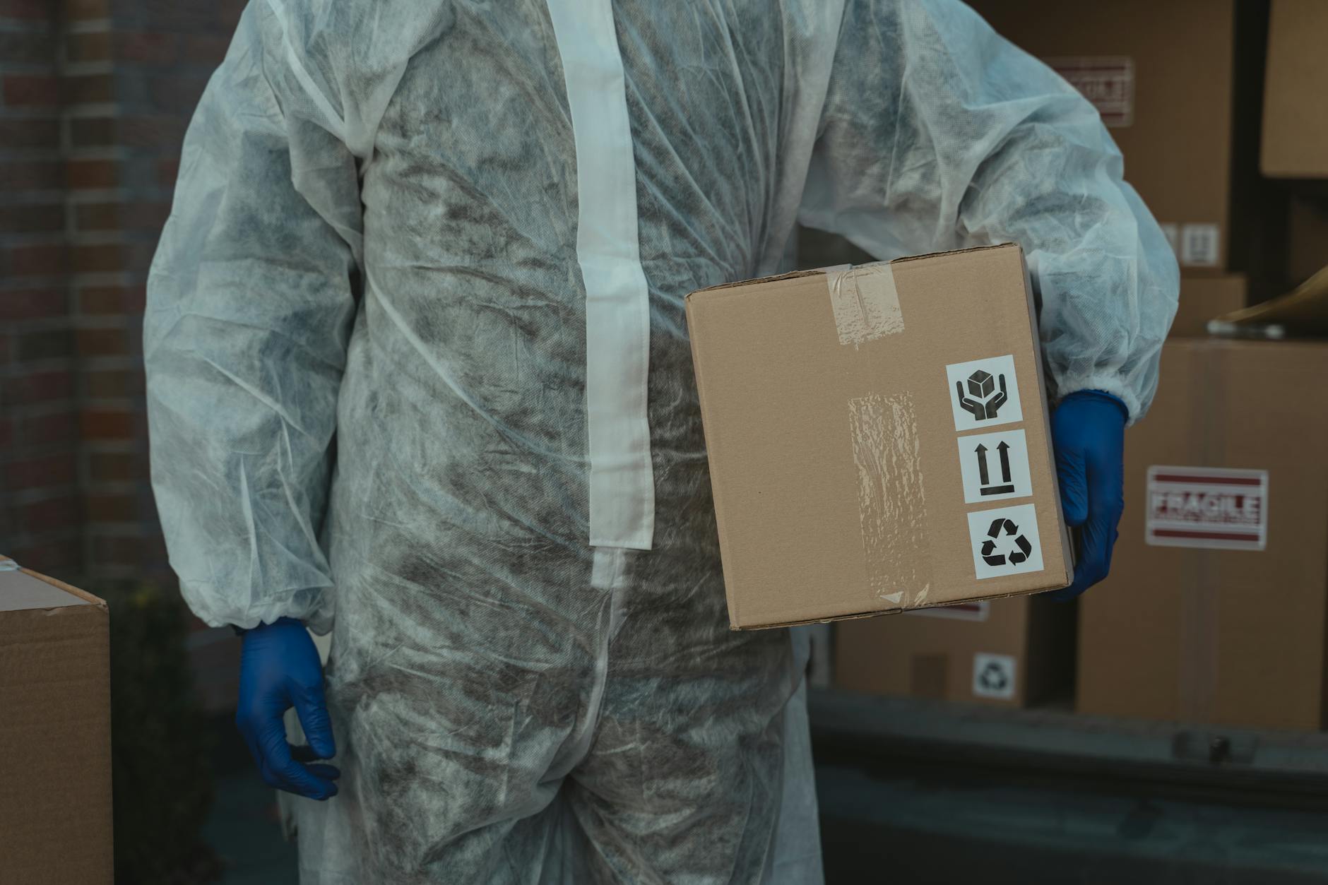 A courier in protective clothing delivers a cardboard package during the day.