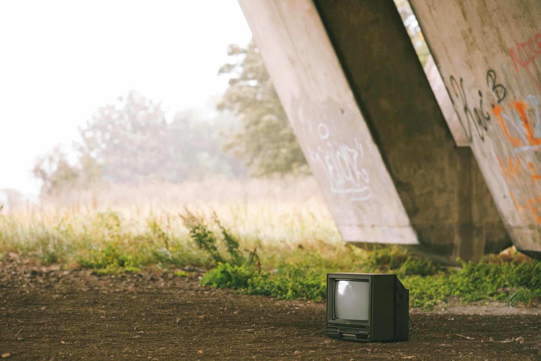 Small black old fashioned television placed on ground near concrete construction with graffiti and grassy terrain with trees on foggy background